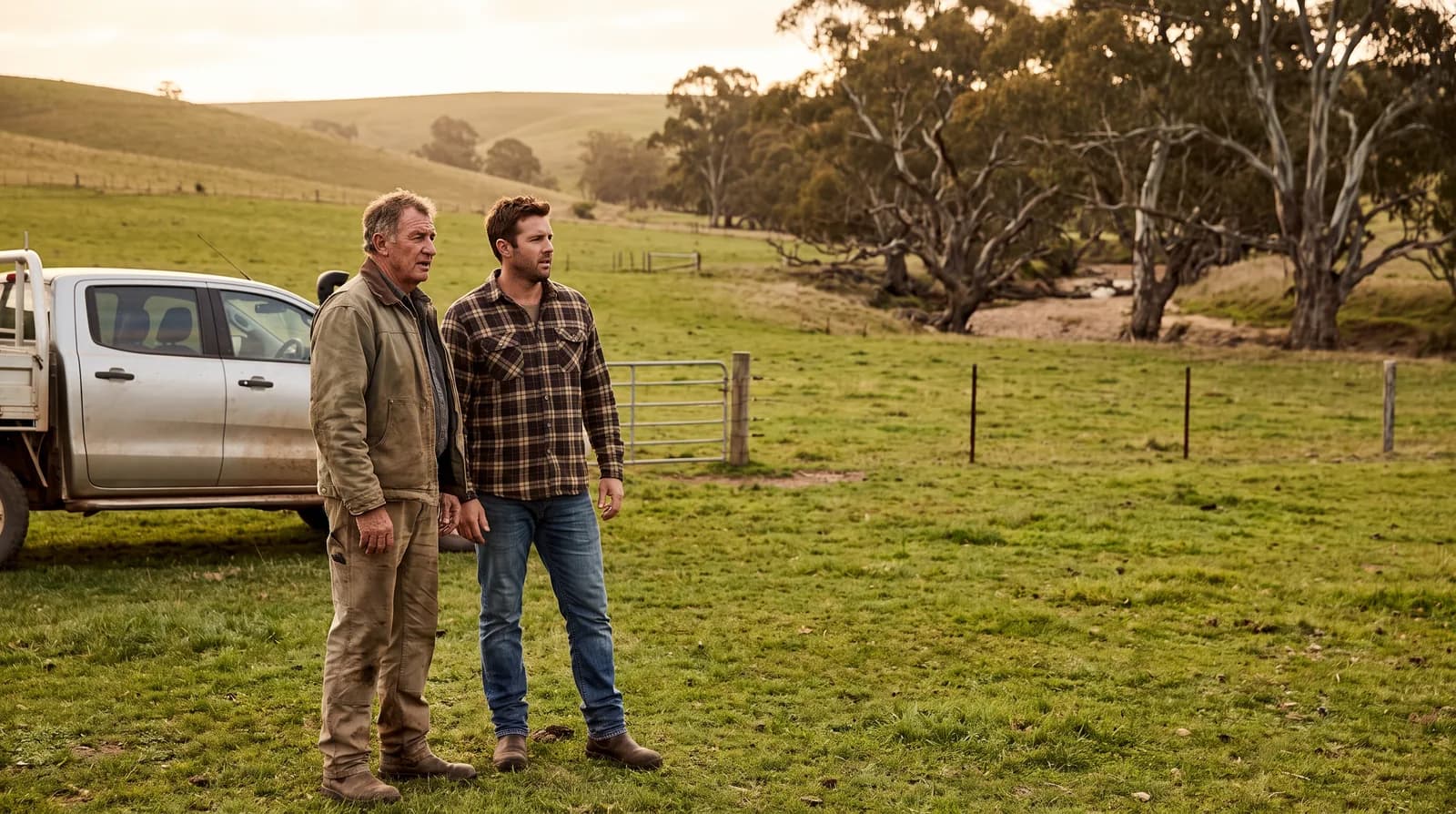 A farmer in his sixties and his adult son standing together in a paddock beside a ute, looking out across the land.