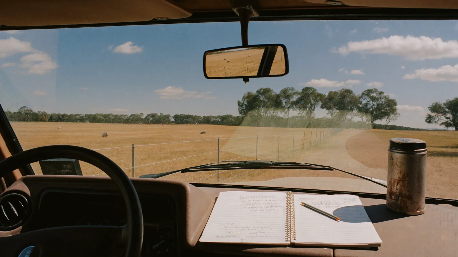 A well-used spiral notebook and pencil on the dashboard of a farm ute, with sunlit paddocks, a fence line and gum trees through the windscreen.