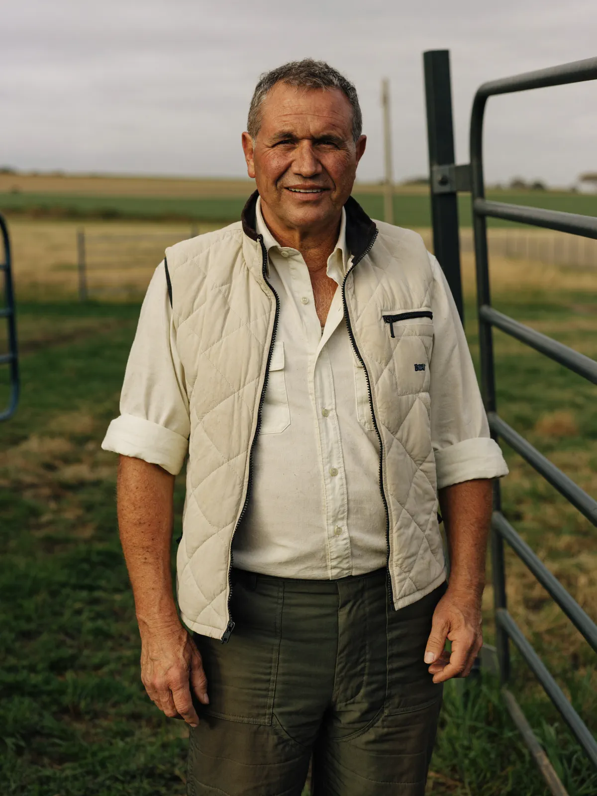 Chris, a farm business consultant in his late forties in a work shirt and quilted vest, standing at a farm gate with green paddocks behind him.