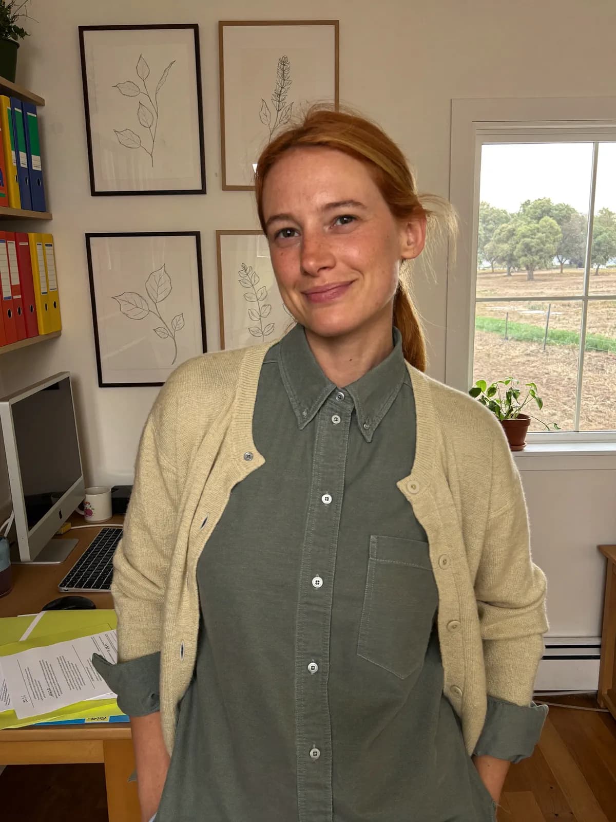 Denise, a financial planner in her late thirties with strawberry blonde hair tied back, wearing a sage shirt and a cream cardigan, standing in her small country-town office with framed botanical drawings on the wall behind her and a fenced paddock through the window.
