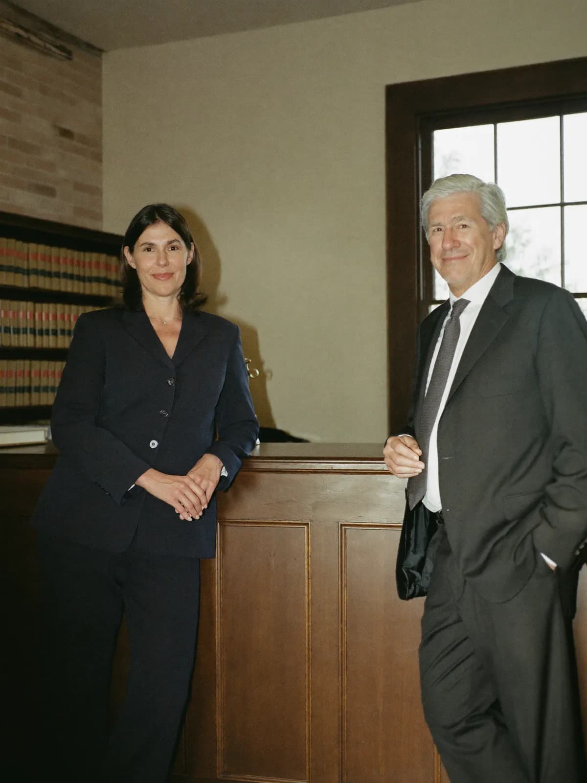 Heather and Peter, the two principals of a regional law firm, standing apart at the counter of an old country law office lined with bound legal volumes.