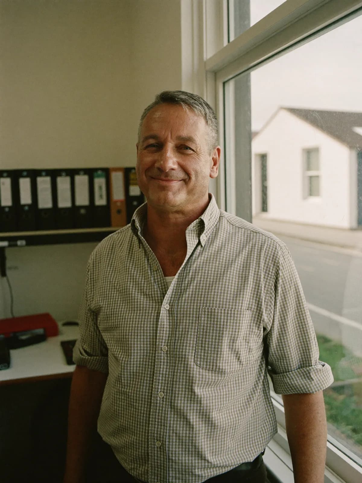 William, an accountant in his mid fifties in a checked shirt with the sleeves rolled, standing by the window of a small country-town accounting office.