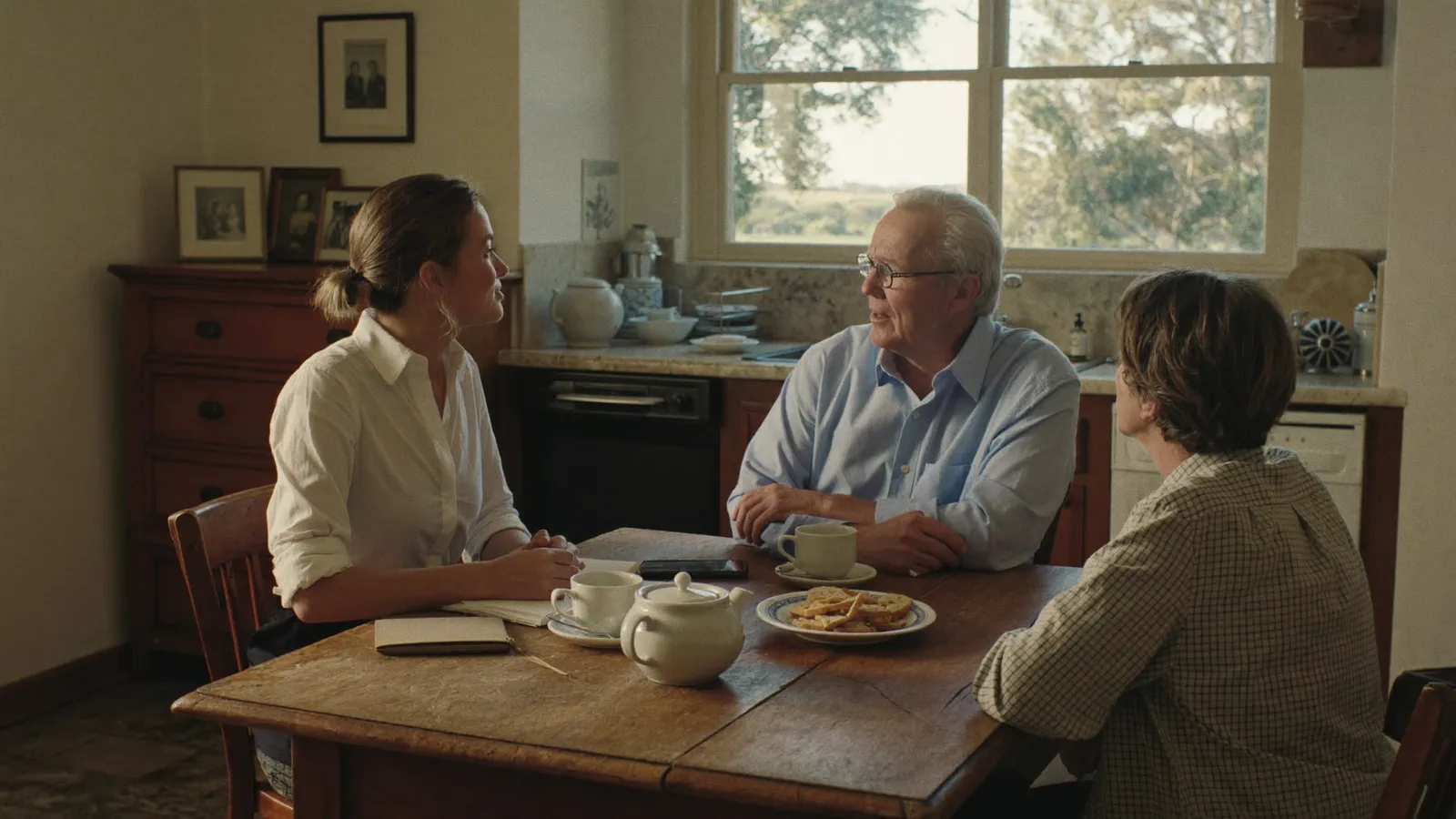 A professional adviser sitting at a farmhouse kitchen table with an older farming couple in warm afternoon light, tea and biscuits on the table and her notebook and phone set to one side.